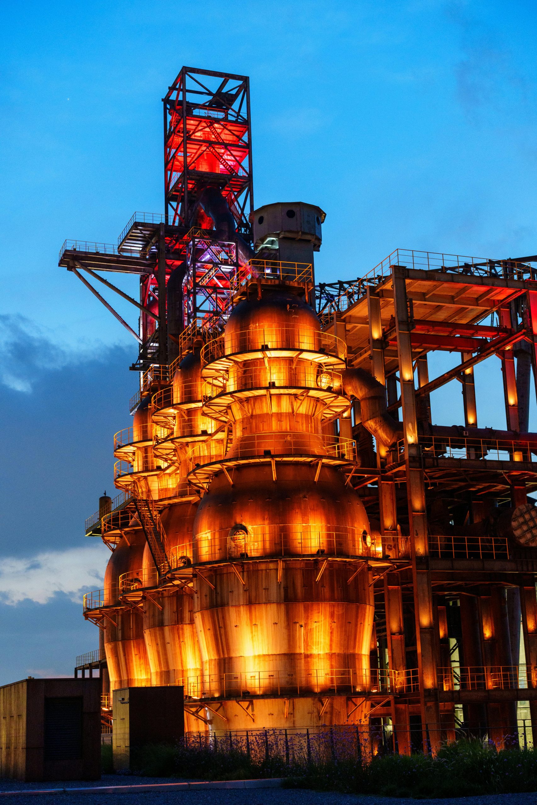 A brightly lit industrial structure stands against a twilight sky, showcasing vibrant colors and intricate design.